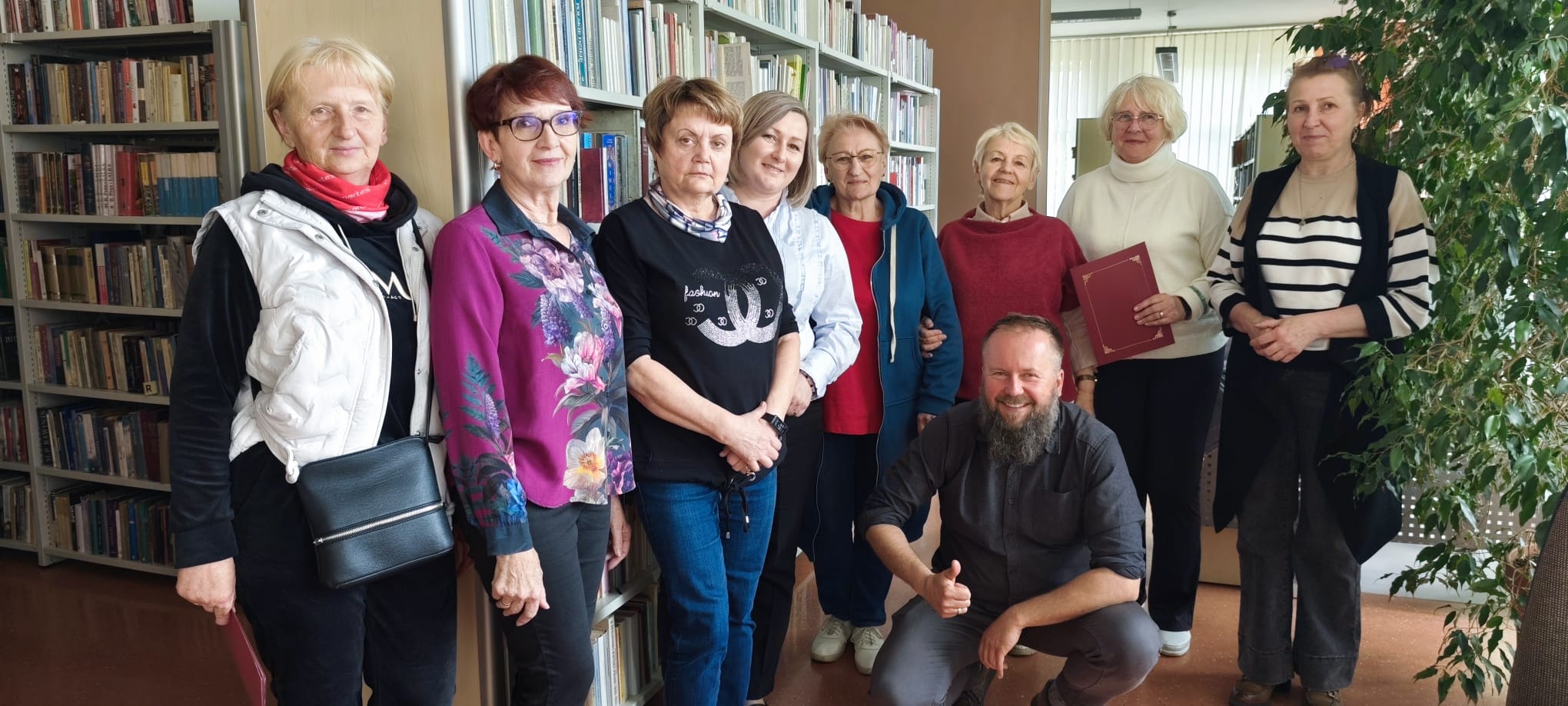Pamiątkowe zdjęcie grupy os&oacute;b w&nbsp;bibliotece.  Opis szczeg&oacute;łowy: Zdjęcie przedstawia dziewięć os&oacute;b pozujących wsp&oacute;lnie w&nbsp;otoczeniu regał&oacute;w z&nbsp;książkami.  Postacie: * Wzdłuż regał&oacute;w po lewej stronie stoi siedem kobiet ubranych w&nbsp;stroje codzienne i&nbsp;wizytowe (m.in. biała kamizelka, wzorzysta koszula, czarna bluza). Jedna z&nbsp;kobiet w&nbsp;głębi trzyma czerwoną teczkę lub&nbsp;folder.  Po prawej stronie, obok dużej zielonej rośliny doniczkowej, stoi &oacute;sma kobieta w&nbsp;pasiastej bluzce i&nbsp;ciemnej kamizelce.  Na pierwszym planie, w&nbsp;środkowej części, kuca mężczyzna z&nbsp;brodą, ubrany w&nbsp;ciemną koszulę. Uśmiecha się i&nbsp;pokazuje gest kciuka w&nbsp;g&oacute;rę.  Tło: * Scena rozgrywa się w&nbsp;bibliotece &ndash; za plecami większości os&oacute;b widać wysokie, białe regały szczelnie wypełnione książkami.  Z prawej strony kadr zamyka duża, gęsta roślina doniczkowa.  W tle widać jasną dalszą część pomieszczenia z&nbsp;oknami, co wskazuje na&nbsp;przestronne wnętrze.