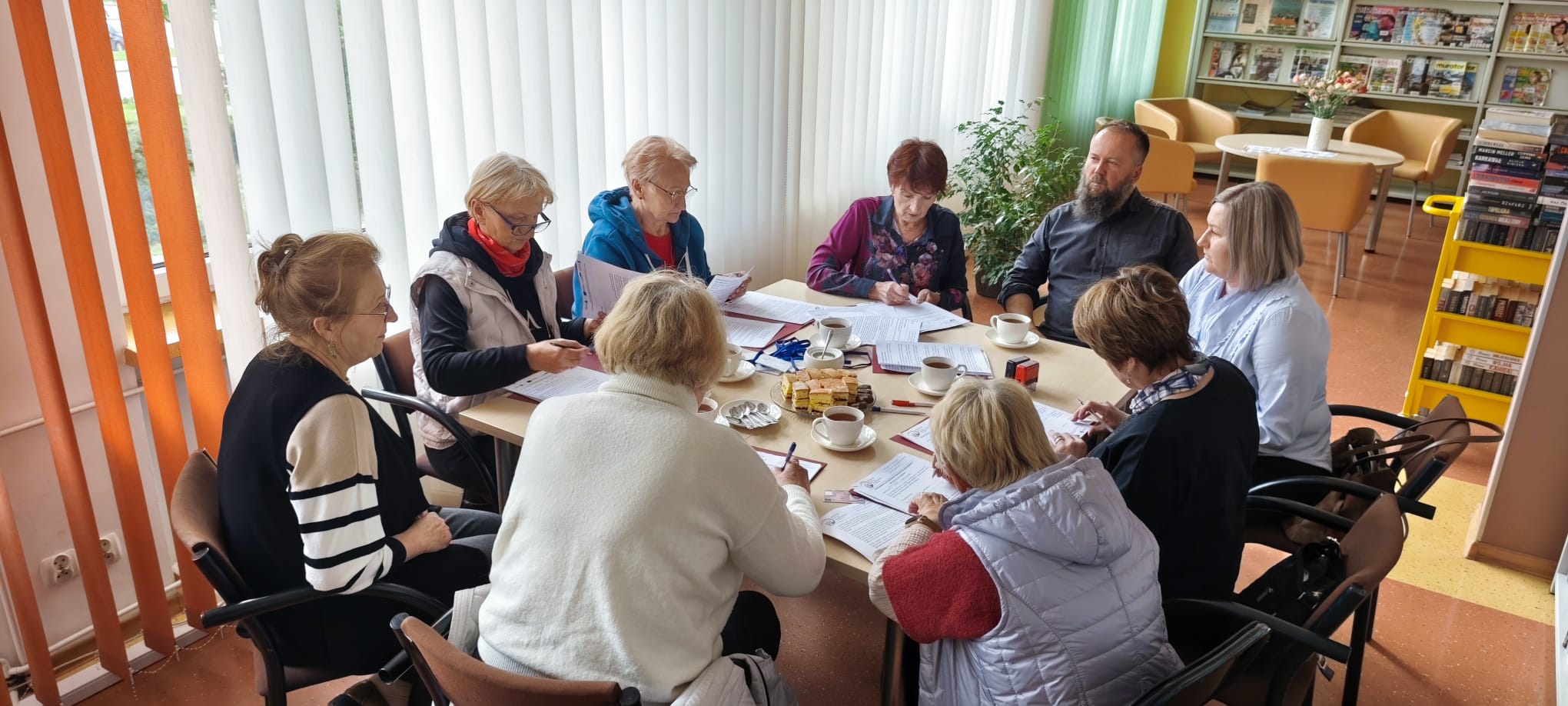 Zdjęcie przedstawia grupę os&oacute;b siedzących przy wsp&oacute;lnym stole w&nbsp;bibliotece podczas pracy z&nbsp;dokumentami.  Opis szczeg&oacute;łowy Czynność: Grupa około dziesięciu os&oacute;b siedzi wok&oacute;ł dużego, jasnego stołu. Uczestnicy są skupieni &ndash; większość z&nbsp;nich przegląda rozłożone arkusze papieru lub&nbsp;składa na&nbsp;nich podpisy.  Uczestnicy: W spotkaniu biorą udział gł&oacute;wnie kobiety w&nbsp;strojach codziennych (swetry, kamizelki, bluzki) oraz&nbsp;jeden mężczyzna z&nbsp;brodą w&nbsp;ciemnej koszuli.  Detale stołu: Na blacie, obok dokument&oacute;w, znajdują się filiżanki z&nbsp;napojami, talerzyk z&nbsp;ciastem, długopisy oraz&nbsp;czerwona pieczątka biurowa.  Otoczenie: * Pomieszczenie jest bardzo jasne dzięki dużym oknom po lewej stronie, przesłoniętym białymi, pionowymi żaluzjami.  W tle widać przestrzeń biblioteczną: regał z&nbsp;książkami i&nbsp;czasopismami oraz&nbsp;strefę relaksu z&nbsp;musztardowymi fotelami i&nbsp;małym stolikiem, na&nbsp;kt&oacute;rym stoją kwiaty w&nbsp;wazonie.  Atmosfera: Scena dokumentuje oficjalne, ale&nbsp;odbywające się w&nbsp;przyjaznej atmosferze spotkanie robocze lub&nbsp;moment podpisywania um&oacute;w/porozumień.