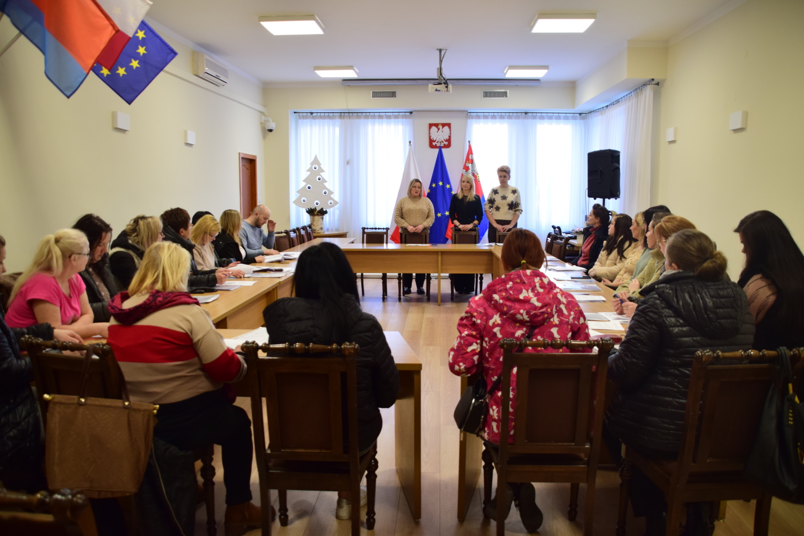 Fotografia dokumentująca spotkanie oficjalne lub&nbsp;szkolenie.  Og&oacute;lny opis: Zdjęcie przedstawia grupę os&oacute;b uczestniczących w&nbsp;spotkaniu w&nbsp;jasnej, przestronnej sali konferencyjnej. Uczestnicy siedzą przy drewnianych stołach ustawionych w&nbsp;kształcie litery &bdquo;U&rdquo;. Panuje atmosfera skupienia, typowa dla oficjalnego wydarzenia lub&nbsp;konsultacji.  Szczeg&oacute;ły kompozycji:      Pierwszy plan: Widoczne są plecy i&nbsp;profile os&oacute;b siedzących najbliżej obiektywu. Uczestnicy są ubrani w&nbsp;stroje codzienne i&nbsp;wierzchnie (kurtki, swetry), co sugeruje okres zimowy. Niekt&oacute;rzy mają przed sobą dokumenty.      Drugi plan (centrum): W głębi sali, przy stole zamykającym układ &bdquo;U&rdquo;, stoją trzy kobiety zwr&oacute;cone w&nbsp;stronę pozostałych uczestnik&oacute;w. Wydają się prowadzić spotkanie lub&nbsp;wygłaszać przem&oacute;wienie.      Tło i&nbsp;symbole: * Za kobietami stojącymi w&nbsp;głębi znajdują się trzy flagi na&nbsp;drzewcach: flaga Polski, flaga Unii Europejskiej oraz&nbsp;flaga regionalna (czerwona z&nbsp;herbem).          Na centralnej ścianie, nad oknami, wisi godło Rzeczypospolitej Polskiej.          Po lewej stronie, w&nbsp;głębi, stoi biała dekoracja w&nbsp;kształcie choinki z&nbsp;ciemnymi ozdobami.          W lewym g&oacute;rnym rogu zdjęcia widoczne są fragmenty flag (polska i&nbsp;unijna) zamocowane na&nbsp;ścianie.
