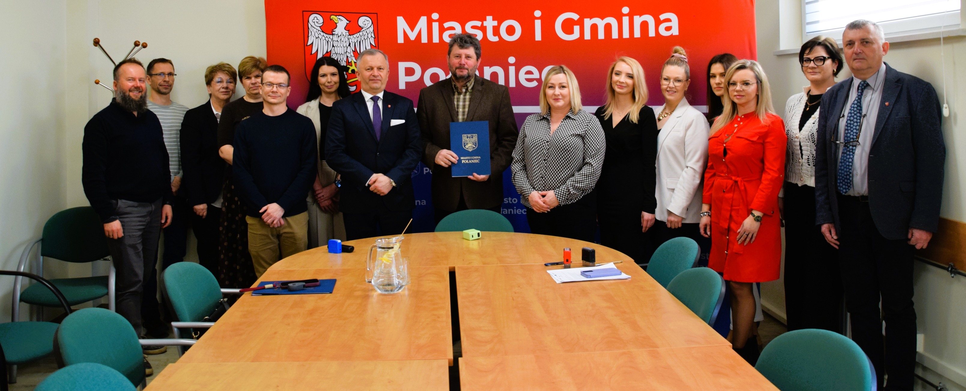 Oficjalne zdjęcie grupowe w&nbsp;sali konferencyjnej na&nbsp;tle baneru &bdquo;Miasto i&nbsp;Gmina Połaniec&rdquo;. Opis szczeg&oacute;łowy      Kompozycja: Na zdjęciu znajduje się grupa około 15 os&oacute;b (kobiet i&nbsp;mężczyzn), stojących ramię w&nbsp;ramię za dużym, drewnianym stołem w&nbsp;kształcie litery U.      Tło: Bezpośrednio za grupą widoczny jest duży, czerwony baner z&nbsp;białym orłem (herbem) oraz&nbsp;białym napisem &bdquo;Miasto i&nbsp;Gmina Połaniec&rdquo;.      Postacie centralne: W środku grupy stoi mężczyzna w&nbsp;brązowej marynarce i&nbsp;koszuli w&nbsp;kratę, kt&oacute;ry trzyma przed sobą niebieską, ozdobną teczkę ze złotym herbem Połańca. Obok niego, po lewej stronie, stoi mężczyzna w&nbsp;ciemnym garniturze i&nbsp;fioletowym krawacie, a&nbsp;po prawej kobieta w&nbsp;czarno-białej, wzorzystej bluzce.      Pozostałe osoby: Pozostali uczestnicy spotkania są ustawieni po obu stronach centralnych postaci. Są ubrani w&nbsp;stroje biznesowe i&nbsp;wizytowe: marynarki, swetry, sukienki (m.in. jedna kobieta w&nbsp;wyrazistej czerwonej sukience) oraz&nbsp;koszule. Wszyscy patrzą w&nbsp;stronę aparatu.      Otoczenie: Sala konferencyjna ma jasne ściany. Wok&oacute;ł stołu ustawione są biurowe krzesła z&nbsp;zielonymi obiciami. Na blacie stołu widać dzbanek z&nbsp;wodą, dokumenty oraz&nbsp;pieczątki biurowe. Po lewej stronie w&nbsp;tle widoczny jest wieszak na&nbsp;ubrania, a&nbsp;po prawej fragment okna.