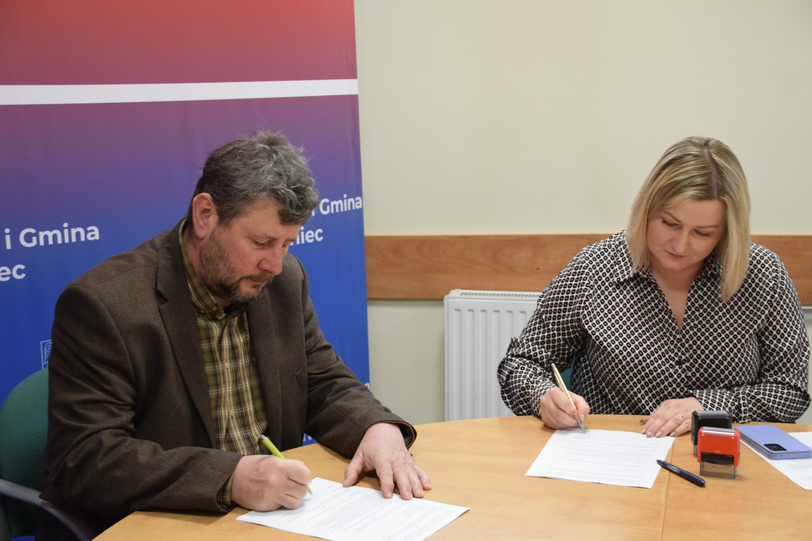 Zdjęcie przedstawiające dwie osoby podpisujące dokumenty przy stole. Opis szczeg&oacute;łowy      Akcja: Fotografia ukazuje moment składania podpis&oacute;w na&nbsp;oficjalnych dokumentach przez kobietę i&nbsp;mężczyznę siedzących przy wsp&oacute;lnym, drewnianym stole.       Osoby:          Po lewej stronie mężczyzna w&nbsp;brązowej marynarce i&nbsp;koszuli w&nbsp;kratę, skupiony na&nbsp;pisaniu zielonym pi&oacute;rem.           Po prawej stronie kobieta w&nbsp;czarno-białej wzorzystej bluzce, podpisująca dokument długopisem.       Otoczenie i&nbsp;detale:          W tle po lewej stronie widoczny jest fragment niebiesko-fioletowego baneru z&nbsp;napisem &bdquo;Miasto i&nbsp;Gmina Połaniec&rdquo;.           Na stole, obok dokument&oacute;w, leżą: niebieski długopis, czerwona pieczątka biurowa oraz&nbsp;smartfon.           Tło za kobietą stanowi jasna ściana z&nbsp;widocznym białym grzejnikiem.