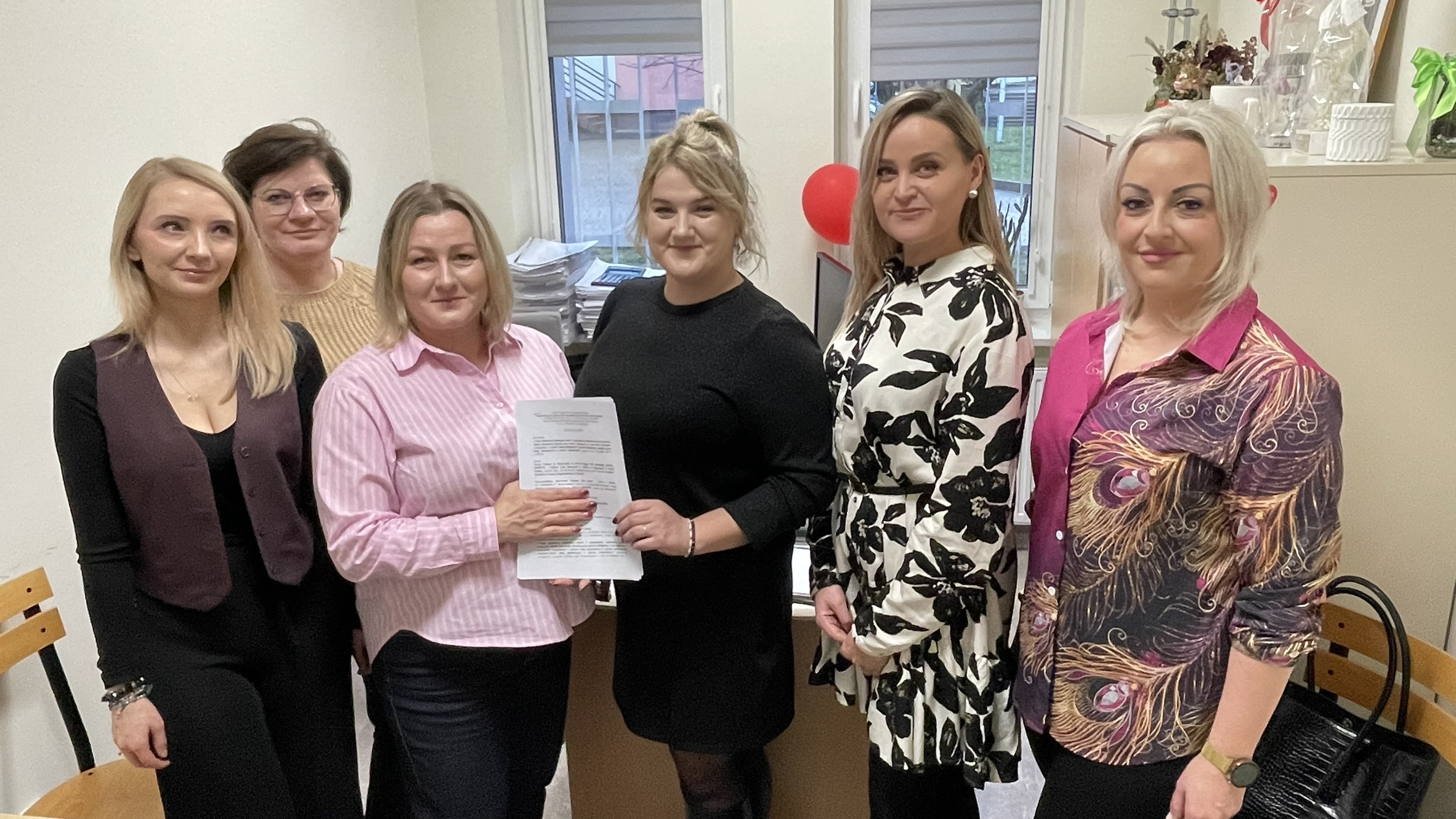 Zdjęcie przedstawia grupę sześciu kobiet w&nbsp;pomieszczeniu biurowym, pozujących do wsp&oacute;lnej fotografii.  Centralny punkt: Dwie kobiety stojące w&nbsp;środku grupy wsp&oacute;lnie trzymają oburącz białą kartkę papieru zadrukowaną tekstem, co sugeruje moment podpisania lub&nbsp;prezentacji oficjalnego dokumentu.  Uczestniczki: Wszystkie kobiety są zwr&oacute;cone twarzą do obiektywu i&nbsp;uśmiechają się. Są ubrane w&nbsp;stroje wizytowe: m.in. czarną sukienkę, r&oacute;żową koszulę, białą bluzkę w&nbsp;czarne wzory roślinne oraz&nbsp;wzorzystą koszulę z&nbsp;motywem pawich pi&oacute;r.  Otoczenie: Tło stanowi jasne biuro. Widoczne są dwa okna z&nbsp;opuszczonymi białymi roletami, biurko z&nbsp;monitorem komputerowym, stosy dokument&oacute;w oraz&nbsp;szafka, na&nbsp;kt&oacute;rej stoi czerwony balon w&nbsp;kształcie serca i&nbsp;ozdobne doniczki.