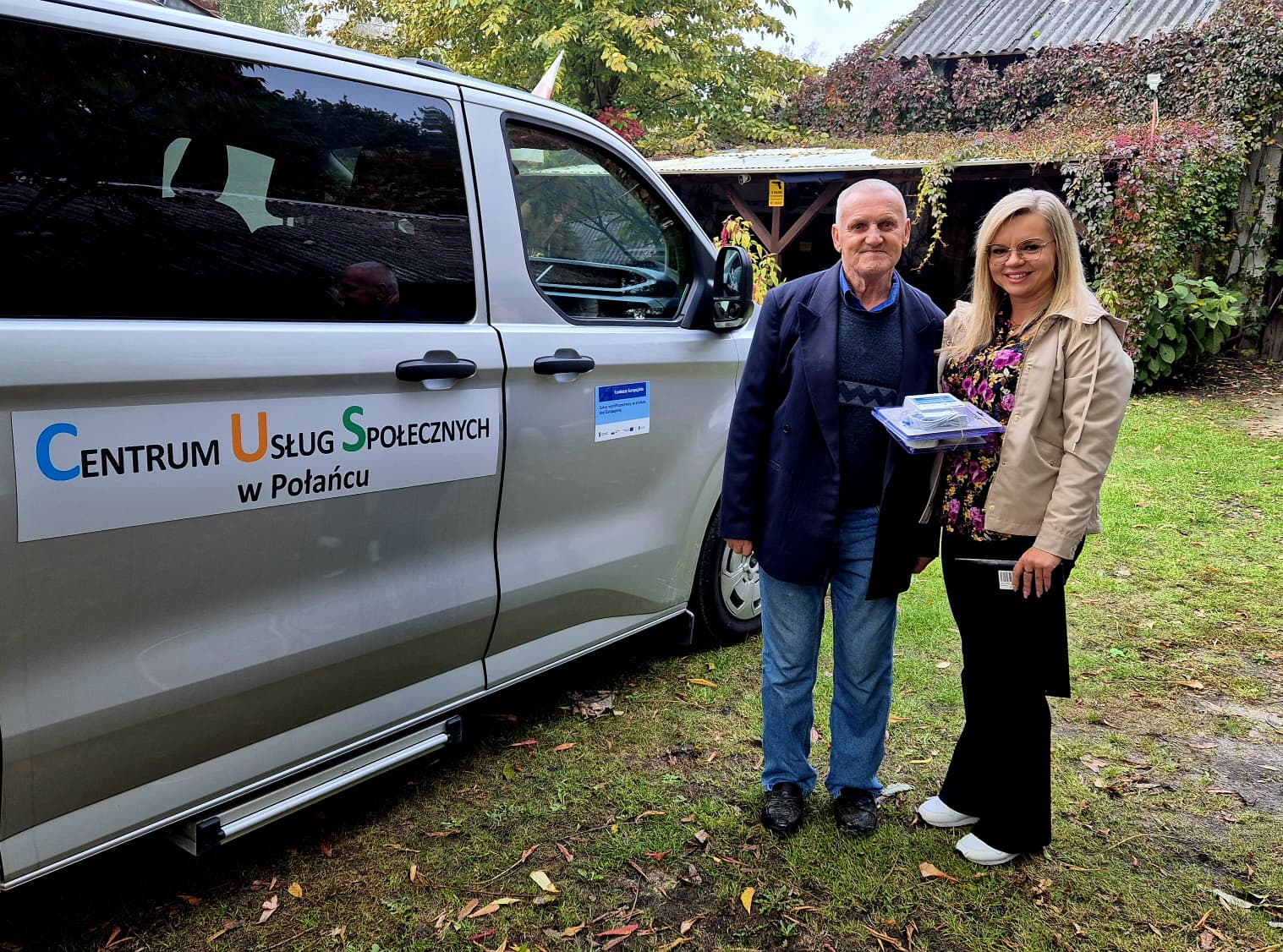 Zdjecie przedstawia dwie osoby, młodszą kobietę i&nbsp;starszego męższczyznę przy mikrobusie z&nbsp;napisem Centrum Usług Społecznych w&nbsp;Połańcu. W tle widać drewnianą stodołę porośniętą bluszczem.