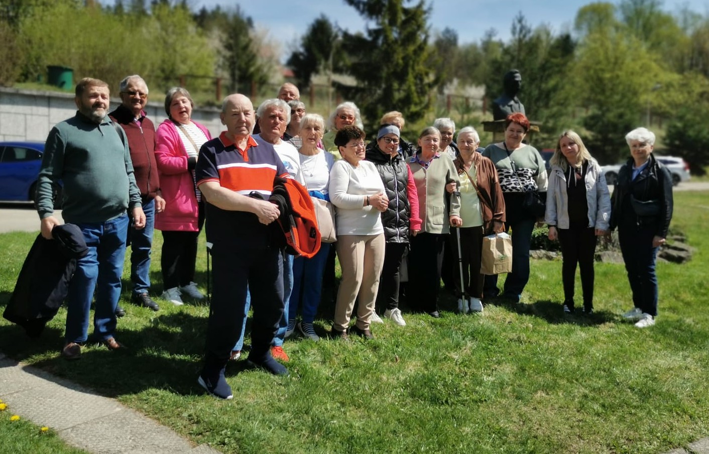 Kompozycja: Zdjęcie przedstawia dużą grupę około 20 os&oacute;b, gł&oacute;wnie senior&oacute;w, pozujących do wsp&oacute;lnej fotografii na&nbsp;zielonym trawniku. Grupa jest ustawiona swobodnie w&nbsp;kilku rzędach.  Uczestnicy: Osoby na&nbsp;zdjęciu są ubrane w&nbsp;kolorową odzież sportową i&nbsp;wycieczkową: bluzy, kurtki, polary oraz&nbsp;wygodne spodnie. Na pierwszym planie wyr&oacute;żnia się mężczyzna w&nbsp;granatowej koszulce polo z&nbsp;szerokim pomarańczowym pasem, trzymający w&nbsp;rękach pomarańczową kurtkę. Niekt&oacute;re osoby wspomagają się laskami do chodzenia.  Tło i&nbsp;otoczenie: * Akcja dzieje się w&nbsp;słoneczny dzień.      W tle po prawej stronie, na&nbsp;podwyższeniu, widoczny jest ciemny posąg (popiersie).      Za grupą rozciąga się pas zieleni z&nbsp;drzewami liściastymi i&nbsp;iglastymi w&nbsp;wiosennej szacie.      Po lewej stronie widoczny jest fragment kamiennego muru oraz&nbsp;zaparkowany niebieski samoch&oacute;d.  Atmosfera: Zdjęcie ma charakter dokumentalny i&nbsp;pogodny; uczestnicy patrzą w&nbsp;stronę obiektywu, dokumentując wsp&oacute;lny wyjazd integracyjny.