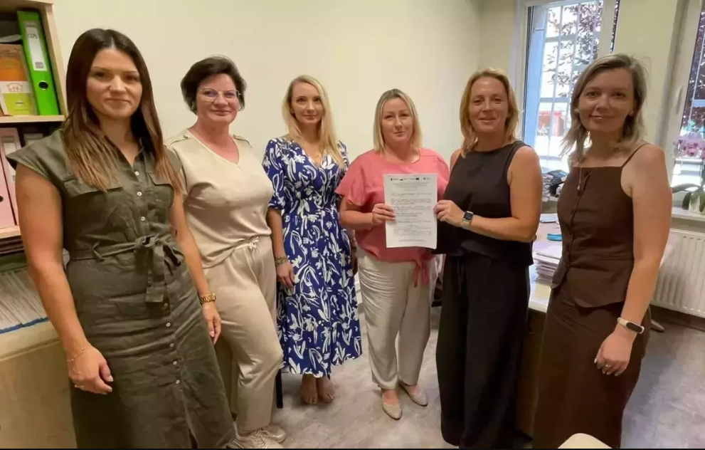 Zdjęcie przedstawia sześć kobiet stojących obok siebie w&nbsp;pomieszczeniu biurowym. Kobieta w&nbsp;r&oacute;żowej bluzce, stojąca w&nbsp;centralnej części, trzyma w&nbsp;dłoniach dokumenty. Wszystkie kobiety są ubrane w&nbsp;r&oacute;żne stroje, od eleganckich sukienek po bardziej swobodne zestawy. W tle widoczne są regały z&nbsp;dokumentami i&nbsp;fragment okna.