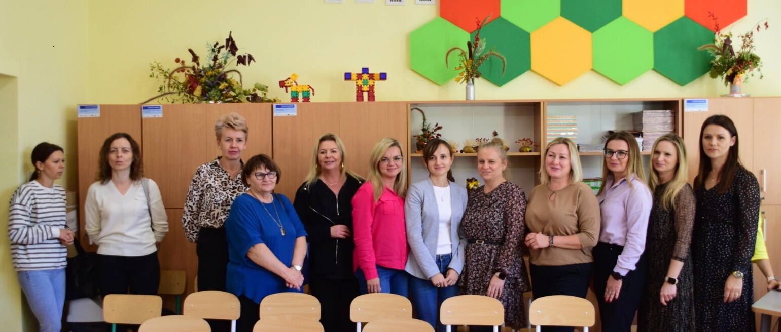 Zdjęcie przedstawia grupę dwunastu kobiet, stojących w&nbsp;dw&oacute;ch rzędach w&nbsp;jasnym pomieszczeniu, przypominającym salę lekcyjną lub&nbsp;świetlicę. W tle widać drewniane szafki, dekoracje ścienne w&nbsp;kształcie kolorowych, geometrycznych plastr&oacute;w miodu (czerwone, pomarańczowe, zielone) oraz&nbsp;p&oacute;łki z&nbsp;książkami. Kobiety są ubrane w&nbsp;r&oacute;żne codzienne stroje i&nbsp;uśmiechają się do obiektywu. Na pierwszym planie widoczne są puste krzesła z&nbsp;oparciami.