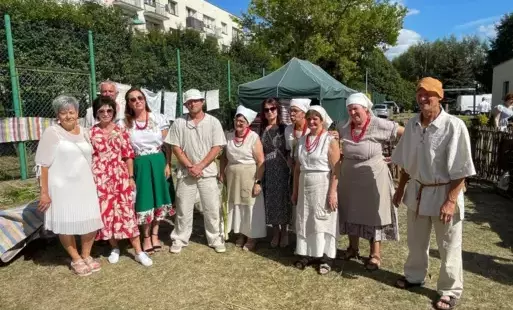 Zdjęcie przedstawia grupę około dziesięciu osób, zarówno kobiet, jak i mężczyzn, stojących na trawniku w słoneczny dzień. Kilka kobiet ubranych jest w tradycyjne, ludowe stroje, z białymi chustami na głowach i czerwonymi koralami. Mężczyźni również mają n