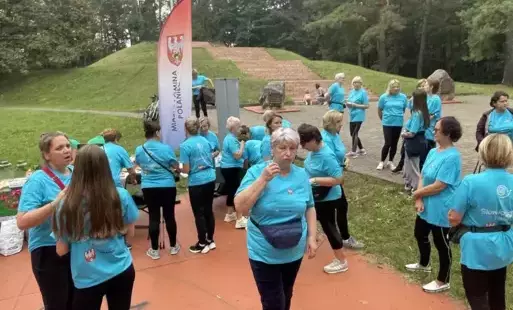 Zdjęcie zrobione z góry, przedstawia grupę ludzi zebranych na utwardzonej ścieżce, otoczonej wysokimi drzewami. Większość osób ubrana jest w jednolite turkusowe koszulki i stoi tyłem do obiektywu, obserwując coś w oddali. W tle, w perspektywie, widać alej