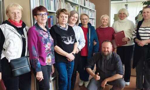Pamiątkowe zdjęcie grupy osób w bibliotece.  Opis szczegółowy: Zdjęcie przedstawia dziewięć osób pozujących wspólnie w otoczeniu regałów z książkami.  Postacie: * Wzdłuż regałów po lewej stronie stoi siedem kobiet ubranych w stroje codzienne i wizytowe (m