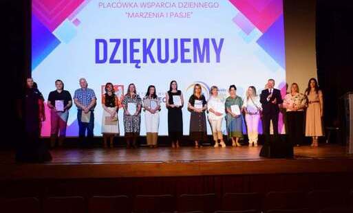 Oficjalne podziękowanie na scenie z udziałem zaproszonych gości i pracowników na tle wielkiego napisu „DZIĘKUJEMY”. Opis szczegółowy      Kompozycja: Szerokie ujęcie przedstawia grupę czternastu osób (kobiet i mężczyzn) stojących ramię w ramię w jednym rz